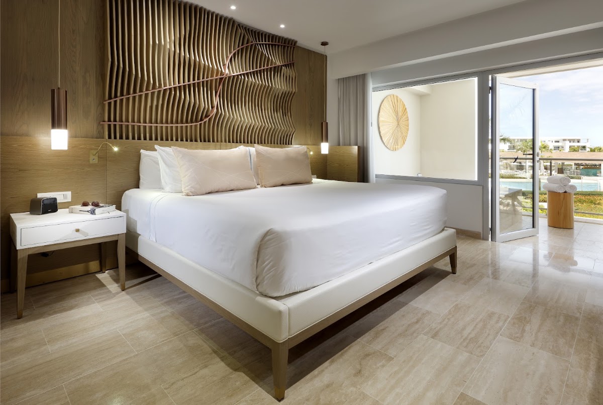 Modern resort bedroom with king bed, wooden headboard wall art, and glass doors opening to a balcony overlooking the pool and grounds.
