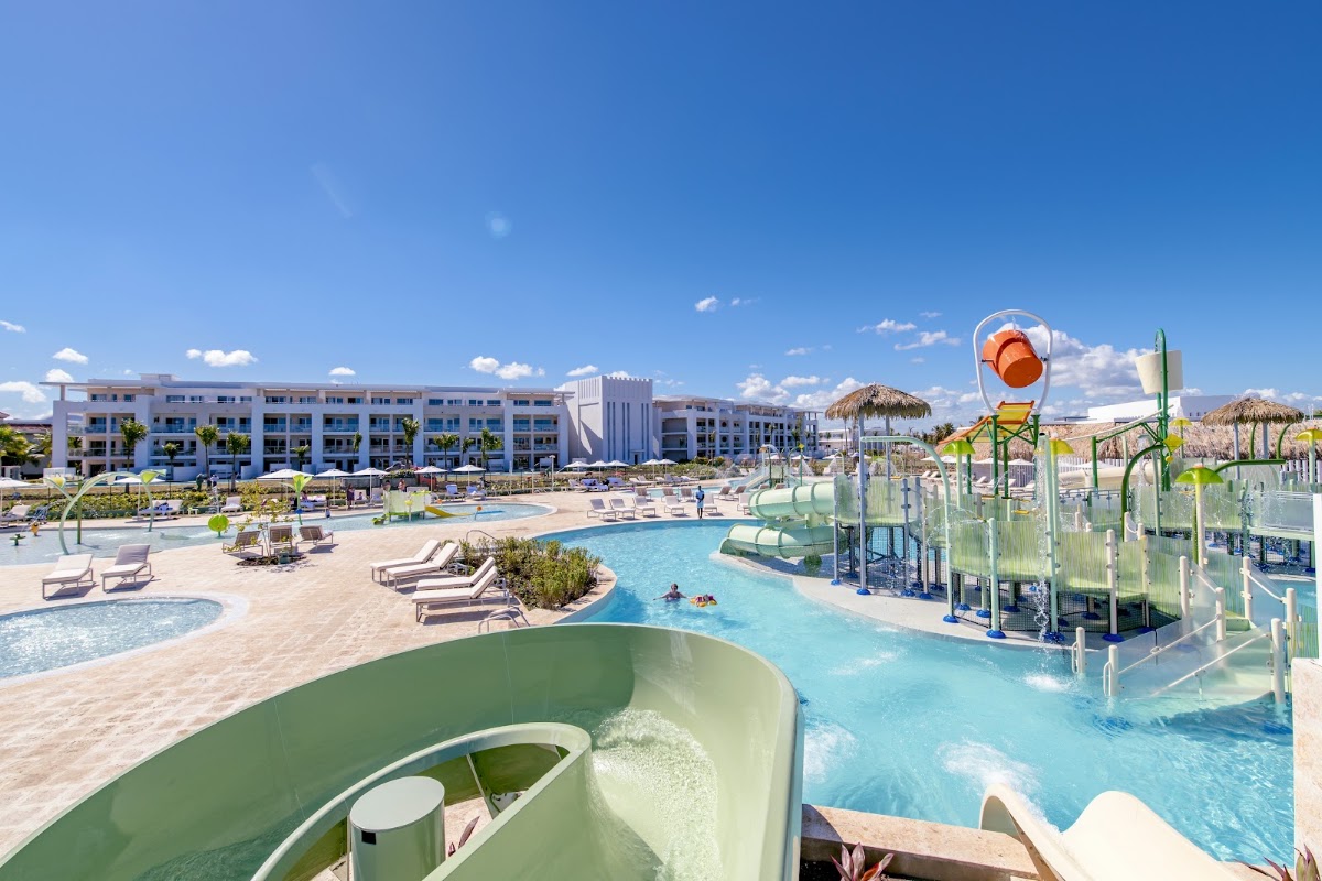 Water slide in foreground with turquoise pool, lounge chairs, and white multi-story resort buildings under clear blue sky.
