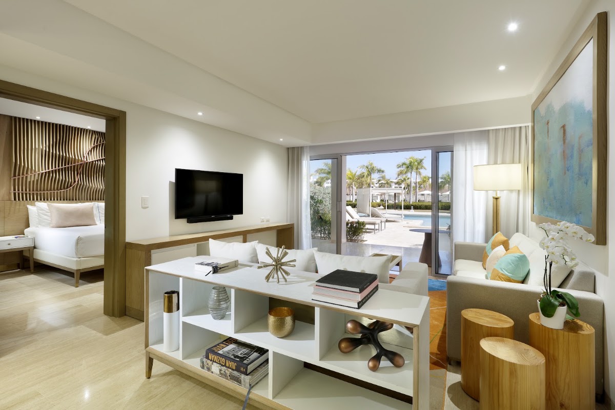 Modern resort suite with living area, bed with wooden frame, TV, white shelving, and glass doors opening to pool and palm trees beyond.