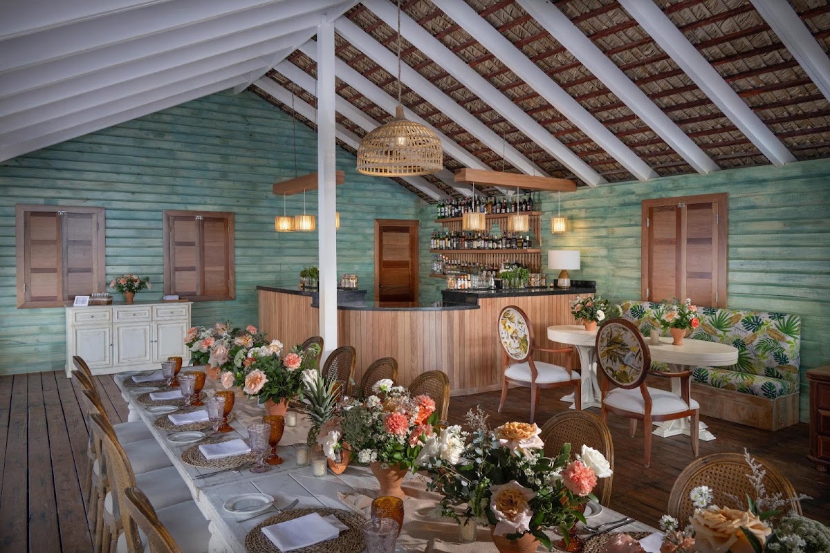 Covered dining pavilion with turquoise shiplap walls, long table set with flowers and place settings, and full bar with woven pendant light overhead.