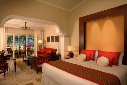 A spacious resort suite with a king bed with red pillows, seating area with red chairs, and ocean-view balcony doors at Paradisus Palma Real Golf & Spa Resort.