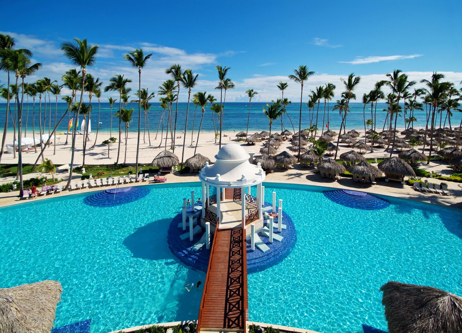 Wooden bridge extending over turquoise pool with white domed structure, palm trees, and sandy beach with thatched umbrellas beyond.