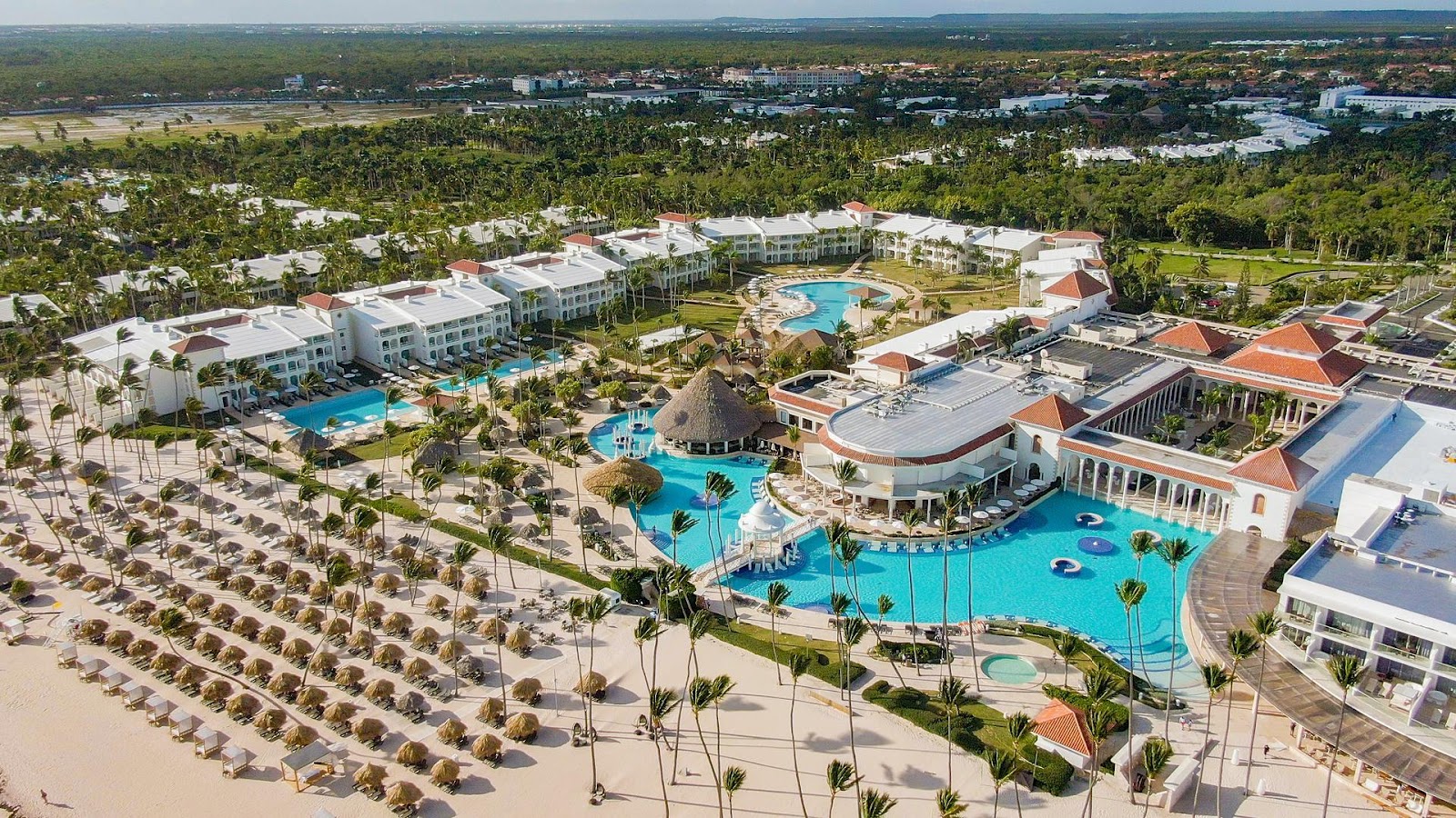 Aerial view of Paradisus Palma Real showing beachfront with palm-lined sand, multiple pools, white buildings, and forested landscape beyond.