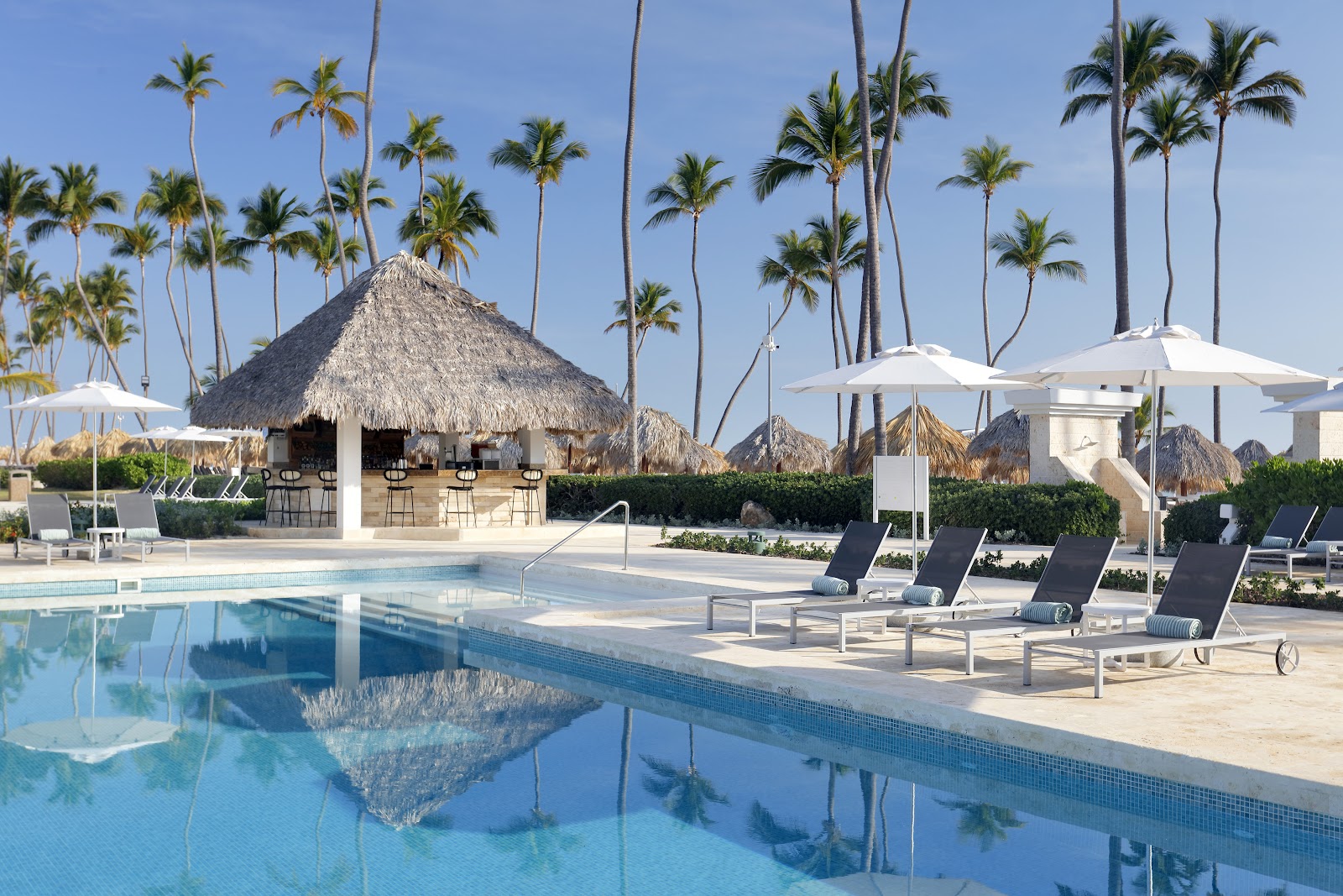 Resort pool with thatched-roof bar and lounge chairs under white umbrellas, backed by tall palm trees and clear blue sky.