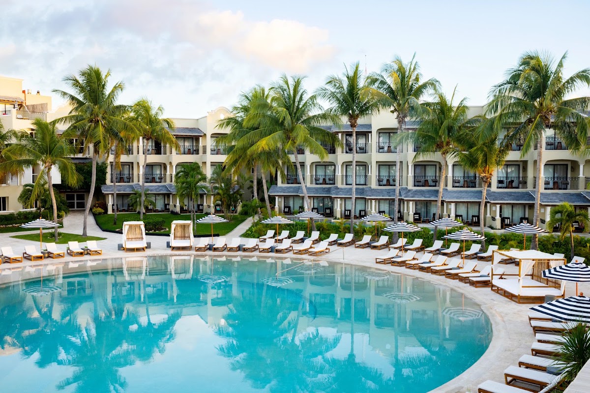 White multi-story resort building with palm trees surrounding a large turquoise pool lined with lounge chairs and umbrellas.