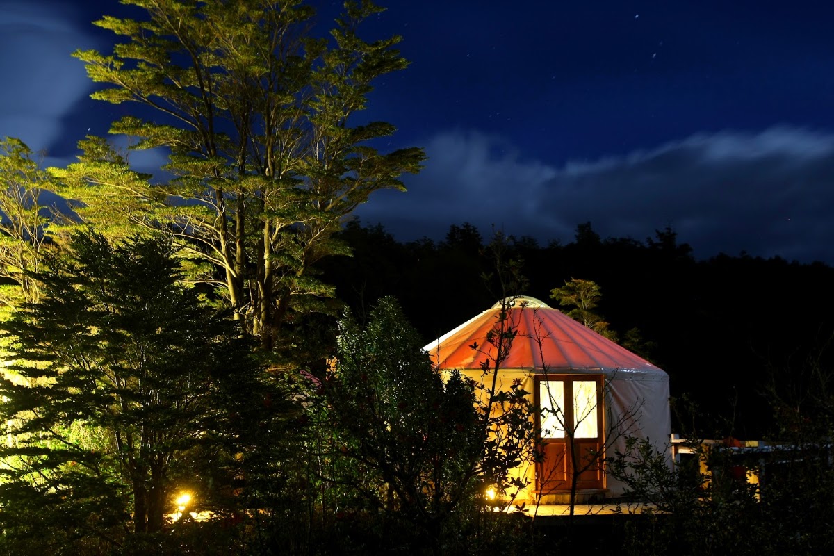 Illuminated yurt with orange conical roof sits among tall trees lit by warm spotlights at dusk at Patagonia Camp.