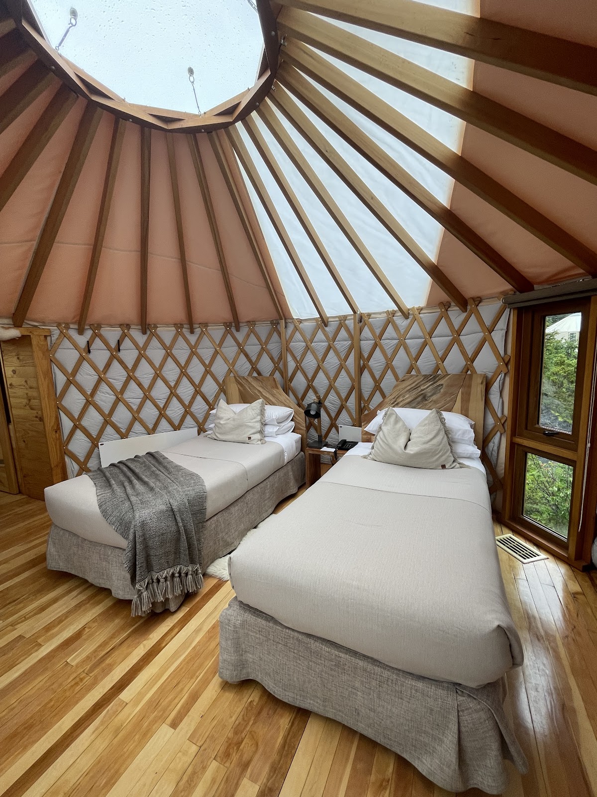 Interior of a yurt bedroom with two beds, wooden lattice walls, tan canvas roof with skylight, and wooden flooring.