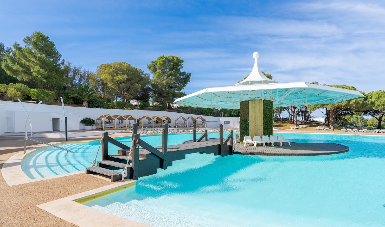 Turquoise pool with wooden bridge, cabanas, and large turquoise umbrella at Pestana Blue Alvor Beach under blue sky.