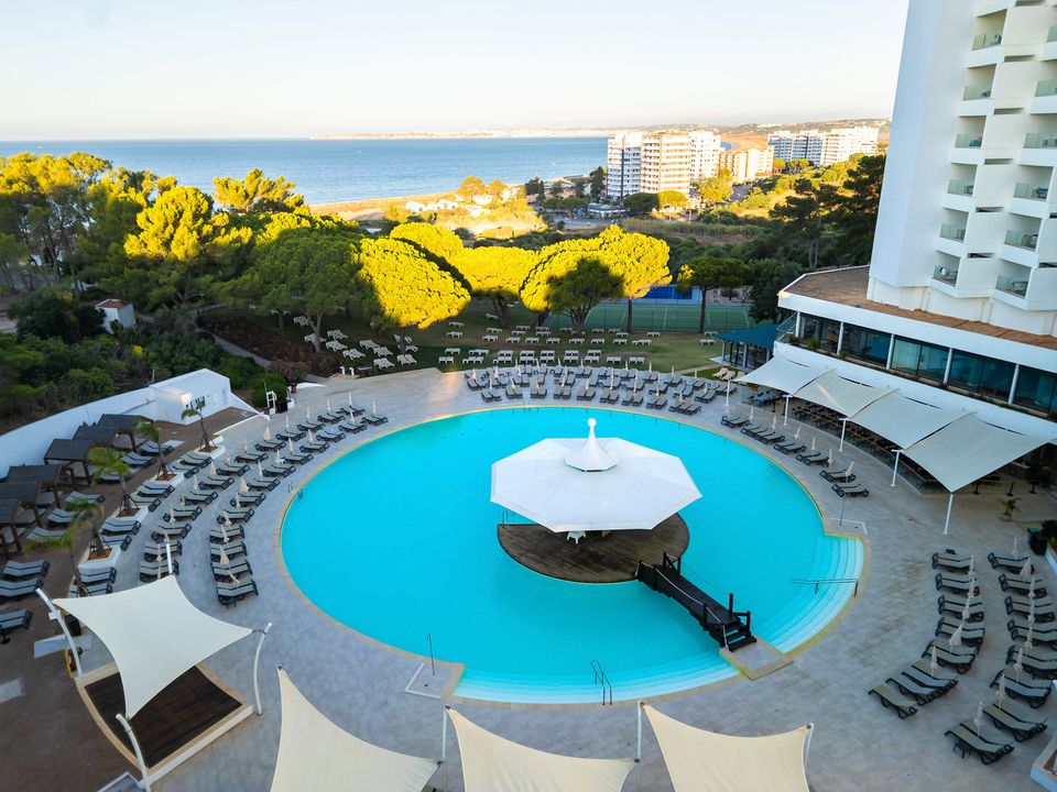 Circular bright blue pool with central white umbrella surrounded by loungers, white resort building, and Mediterranean coastline beyond.