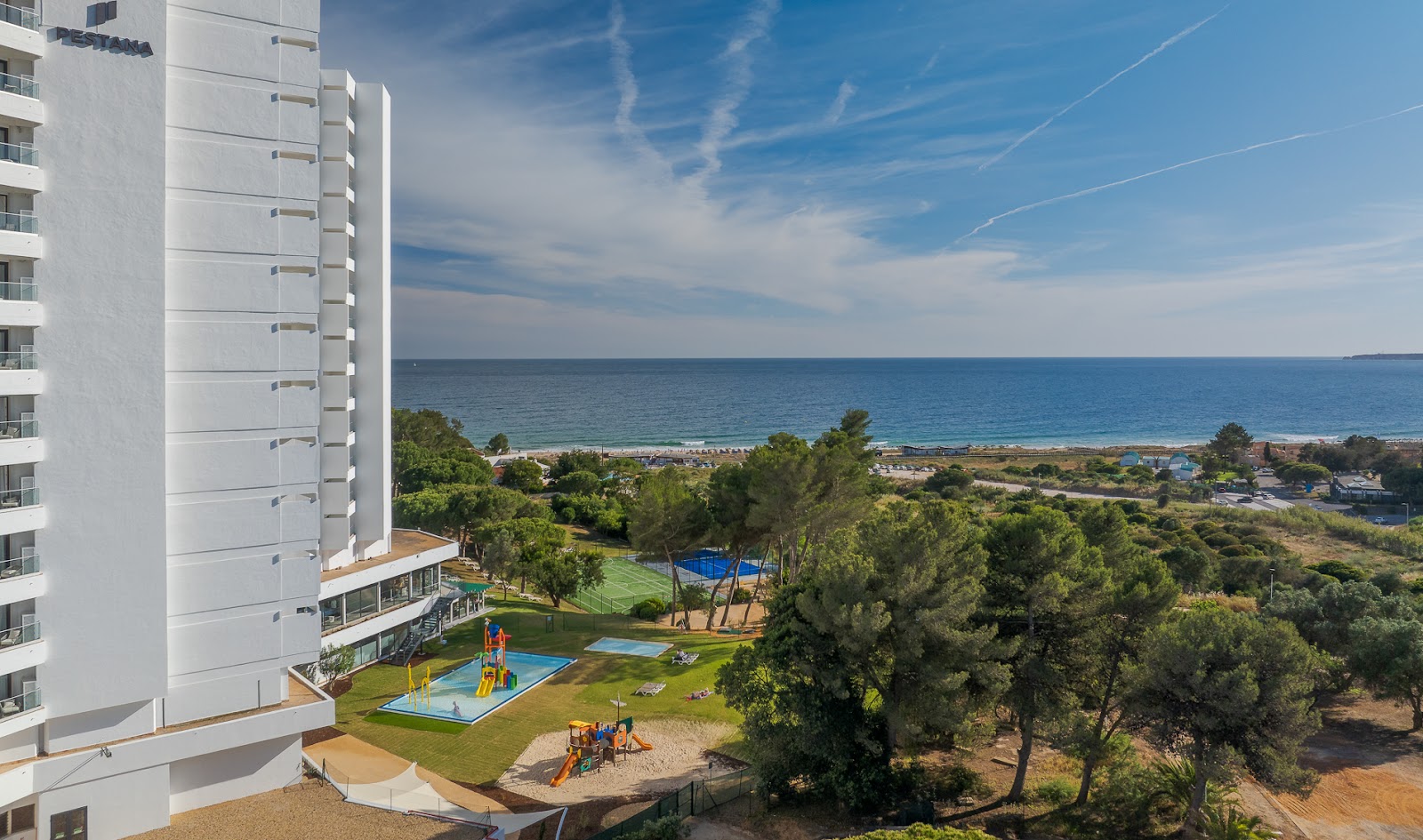 Large turquoise pool with central island and turquoise umbrella at Pestana Blue Alvor Beach, lined with lounge chairs and beach cabanas.