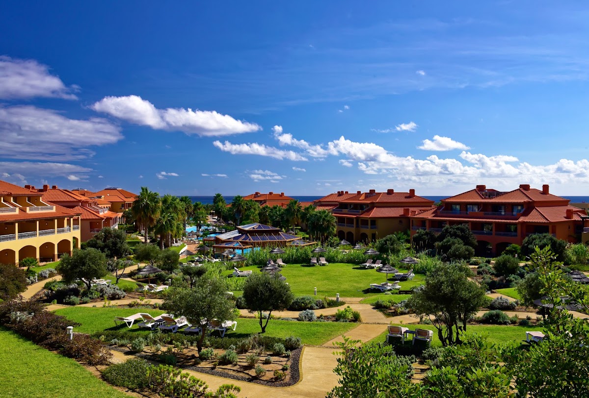 Terracotta-roofed resort buildings overlook manicured grounds with olive trees, lounge chairs, and a pool complex near the ocean under blue sky.