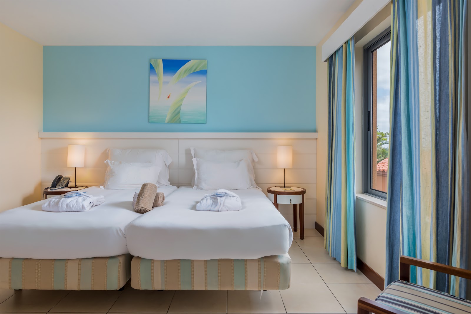 Resort bedroom with two twin beds, white linens, turquoise accent wall, bedside lamps, and sliding glass doors opening to a balcony.