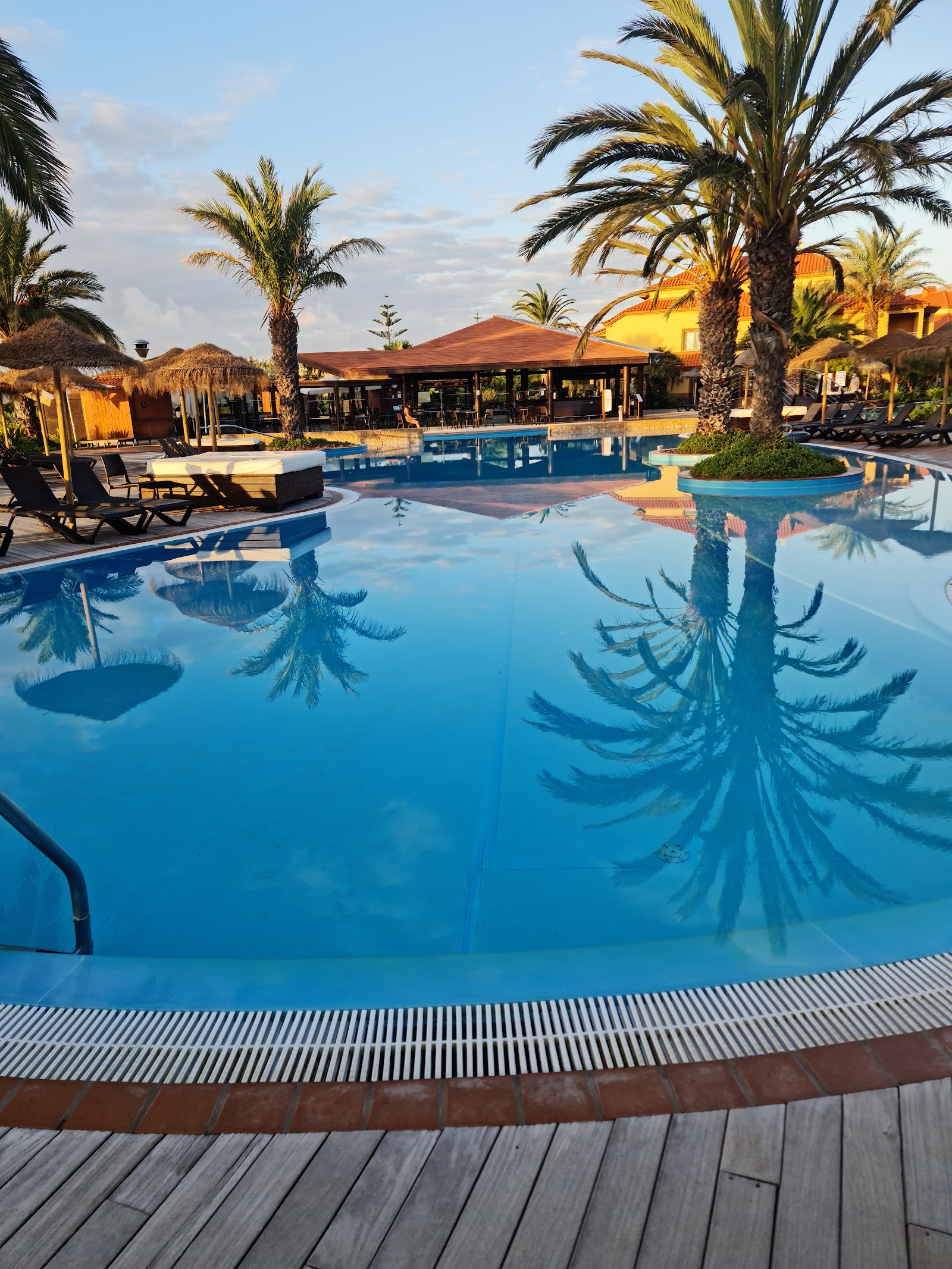 Large blue pool with palm trees and terracotta-roofed resort buildings against hillside landscape under clear sky.