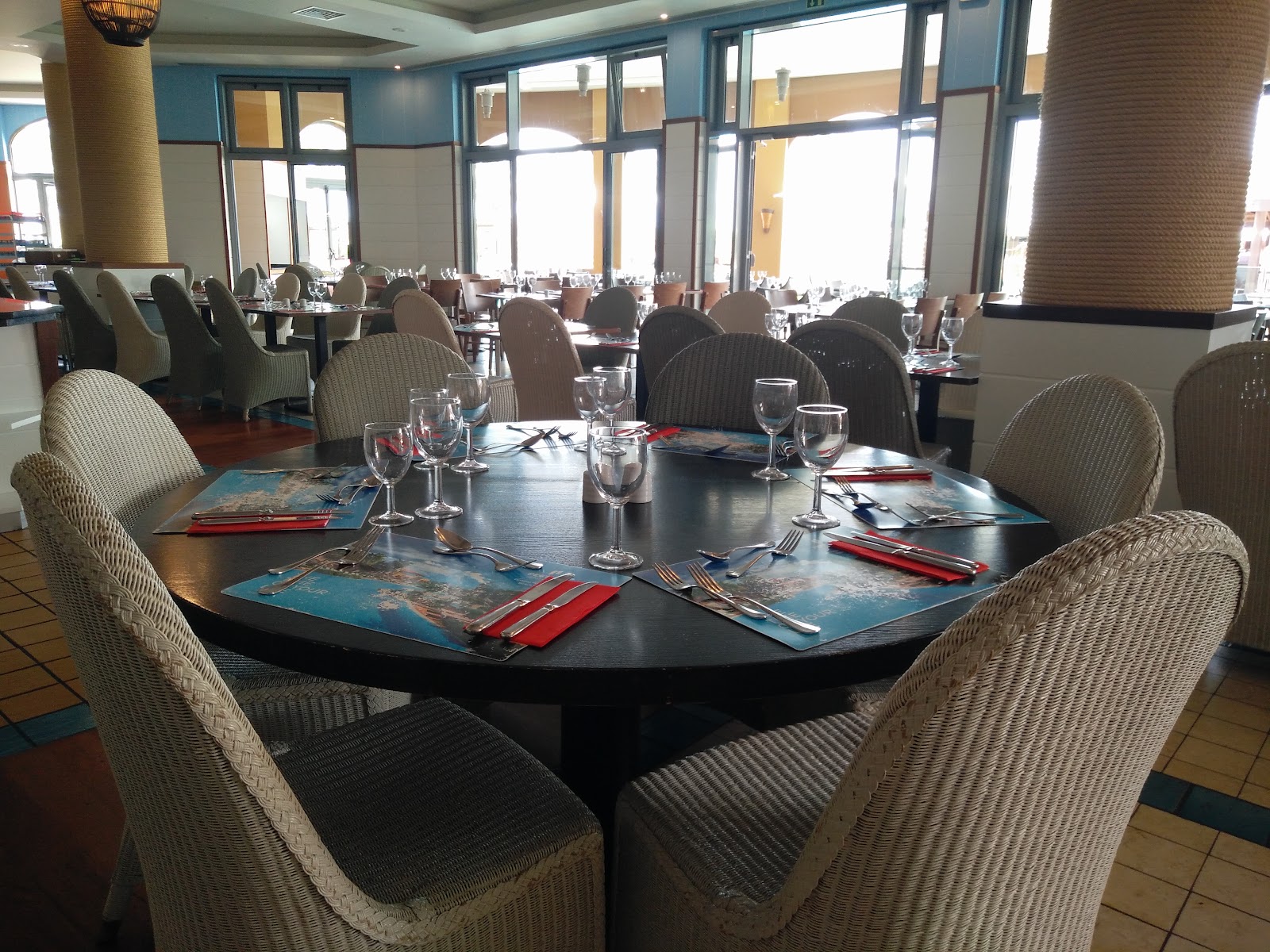 Dining room with round black tables set with placemats, glassware, and red napkins, woven chairs, and large windows overlooking the resort grounds.