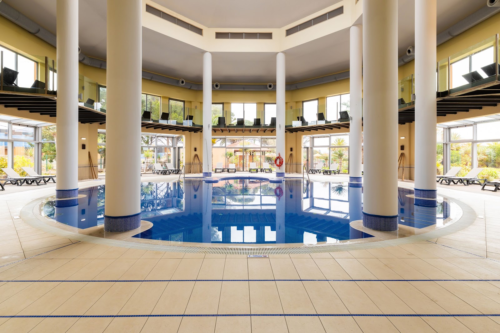 Indoor swimming pool with blue water surrounded by tall white columns, yellow walls, and lounge chairs, at Pestana Porto Santo All Inclusive.