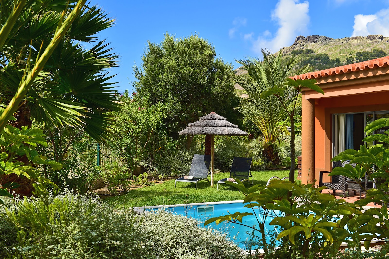 Orange villa with private pool, loungers under thatched umbrella, palm trees and hillside backdrop at Pestana Porto Santo.