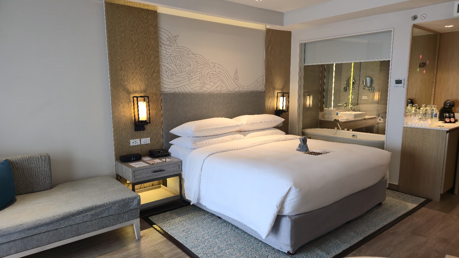 Modern hotel bedroom with king bed, white linens, textured wood headboard with decorative wave artwork, ensuite bathroom visible on right, and gray seating area on left.