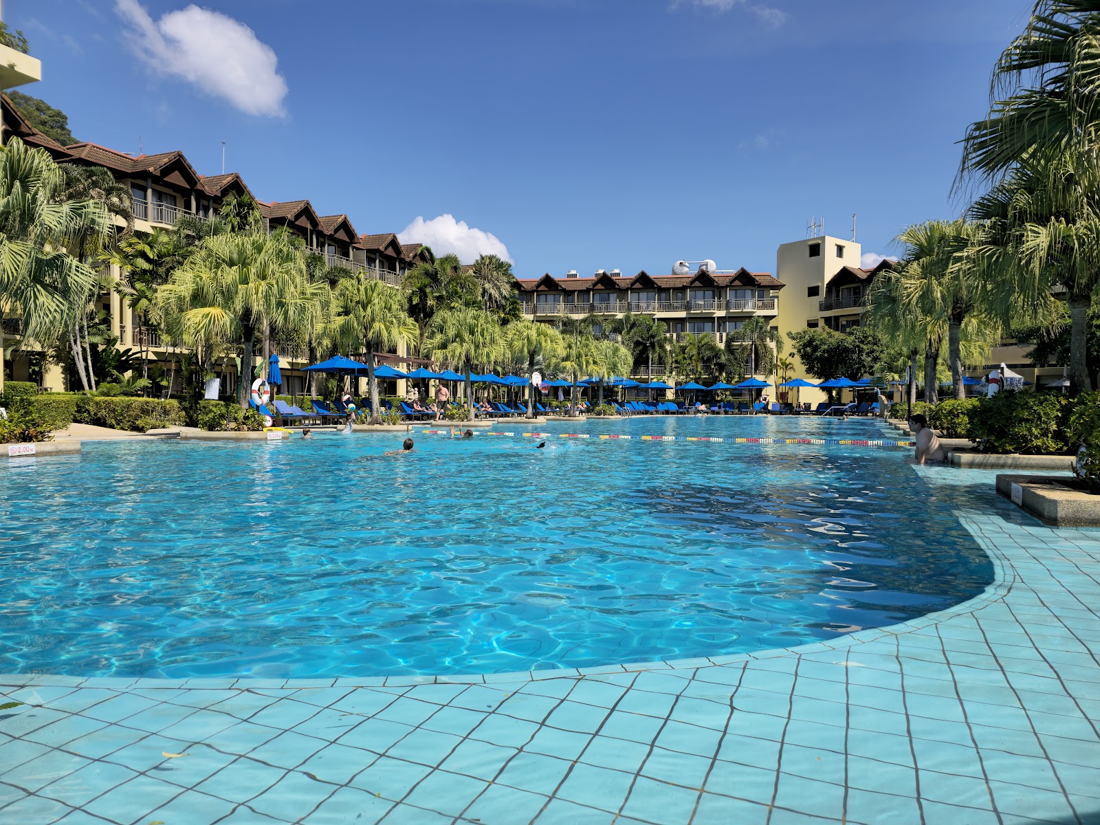 Large resort pool with swimmers, blue umbrellas, and multi-story hotel buildings surrounded by palm trees under clear sky.