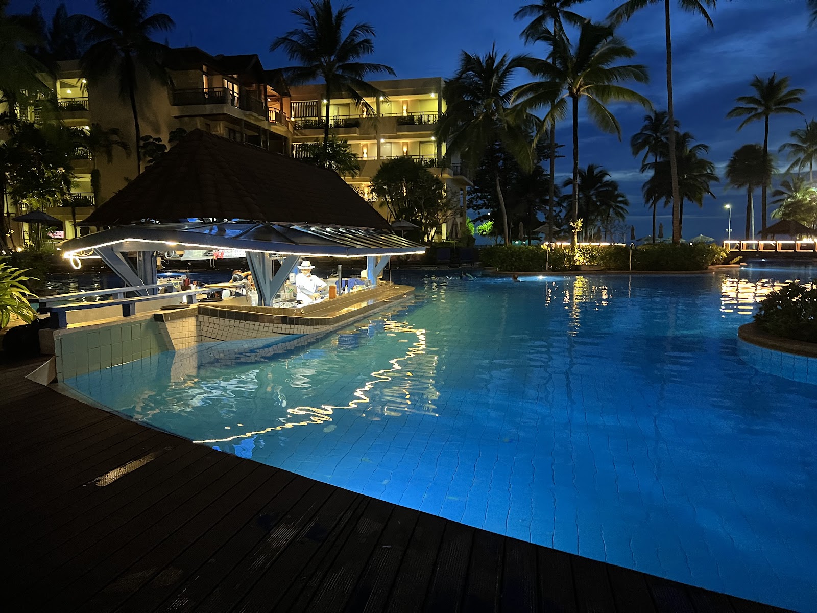 Lit swim-up bar and illuminated resort buildings beside a glowing blue pool at dusk, surrounded by tall palms and ocean view.