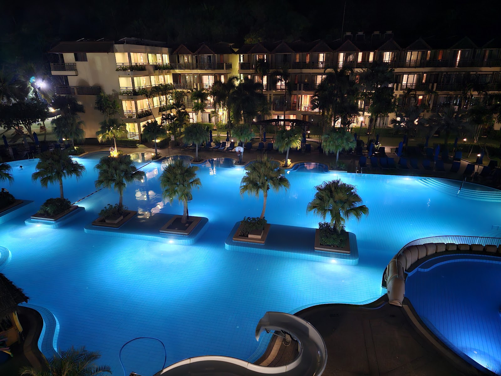 Illuminated lagoon-style pool at night with palm trees, lit resort buildings, and guests relaxing poolside at Phuket Marriott Resort & Spa, Merlin Beach.