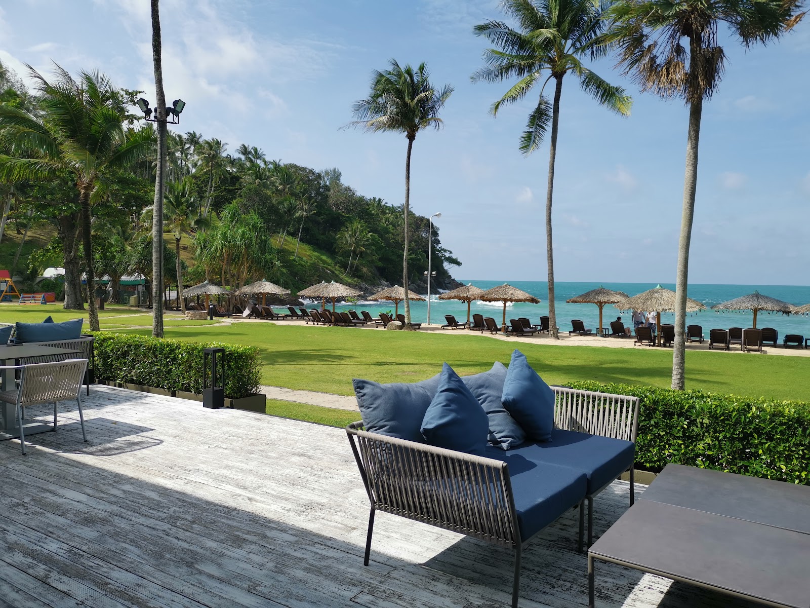 Blue cushioned lounge chair on a wooden deck overlooking the manicured lawn, beach loungers, thatched umbrellas, and turquoise ocean at Phuket Marriott Resort & Spa, Merlin Beach.