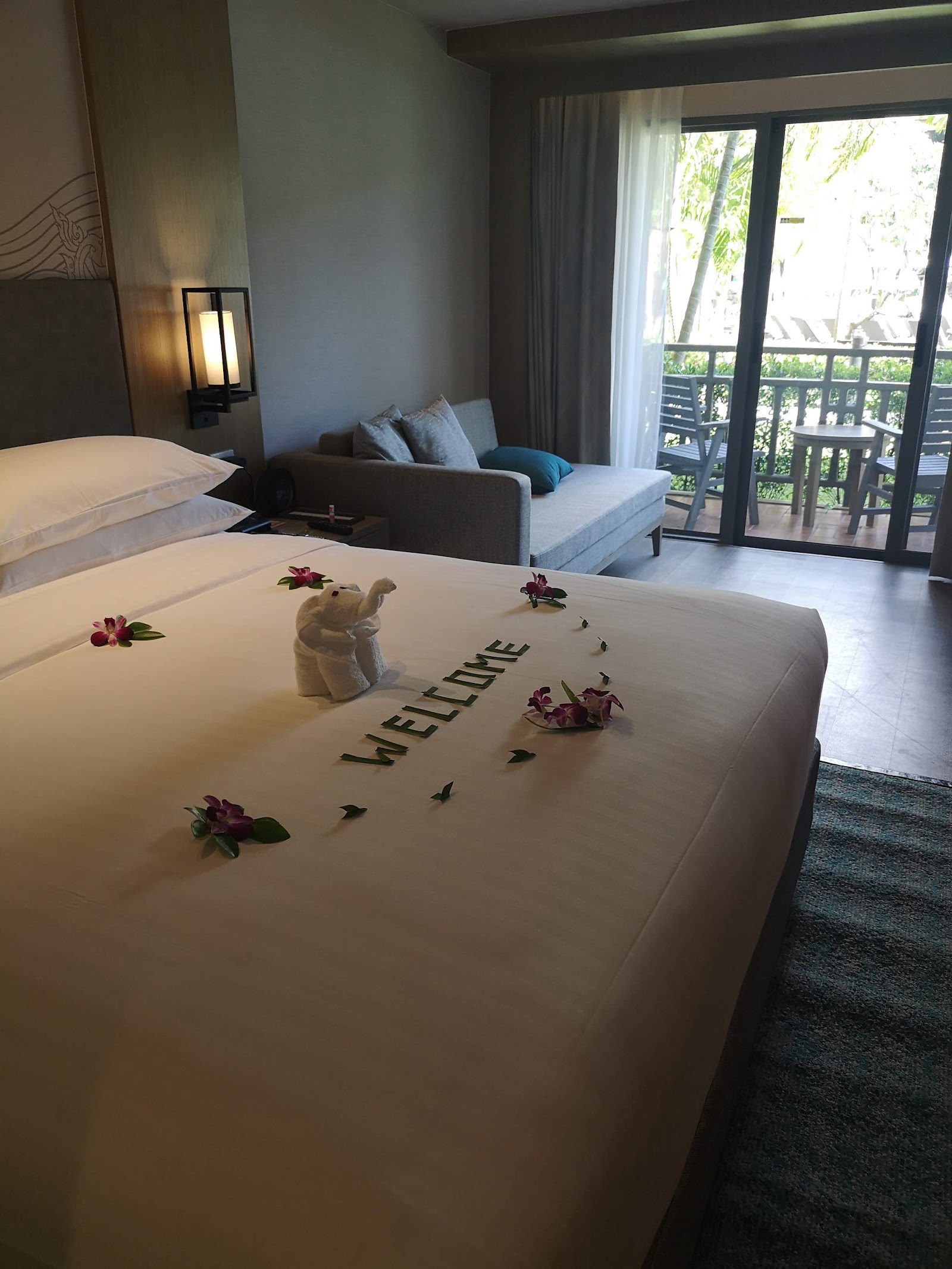 Resort guest room with king bed decorated with welcome message, towel elephant, and red flowers, sofa, and balcony overlooking tropical grounds.