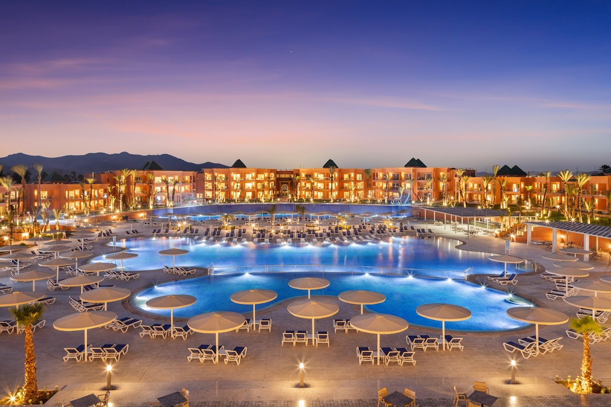 Illuminated pools with tan umbrellas and lounge chairs at Pickalbatros Sungo Club, with ochre buildings and mountains at dusk.