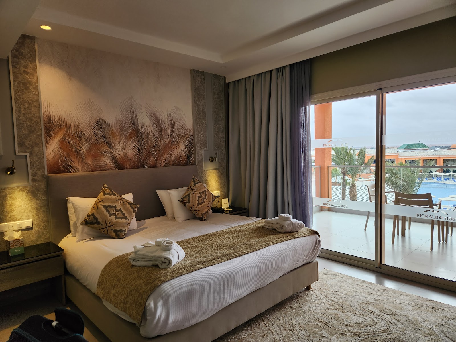 Hotel bedroom with terrace doors framing a resort pool, palm trees, and clay-colored buildings under clear sky.
