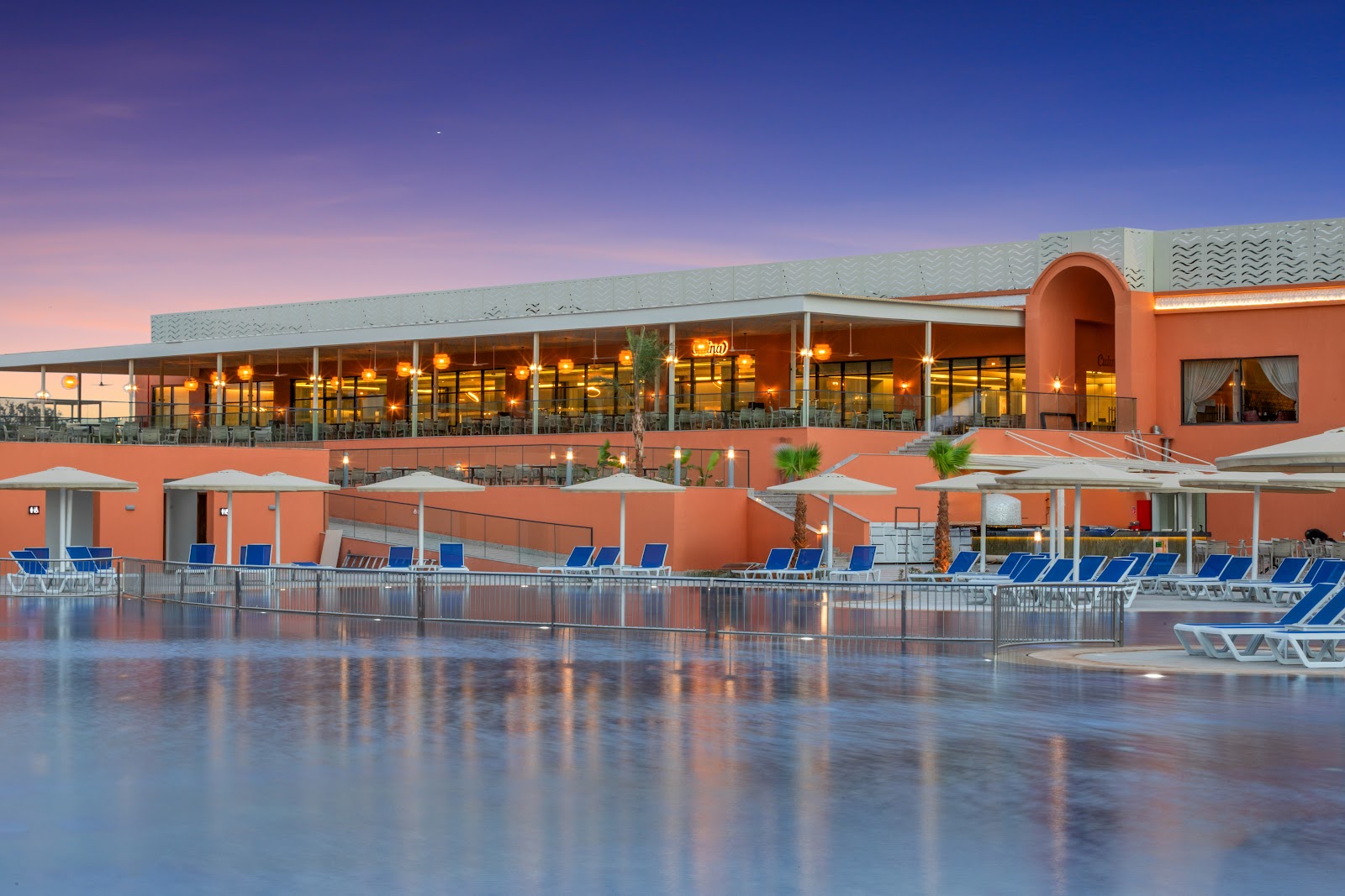 Twilight view of the pool deck with loungers, shaded pergolas, and an orange Moroccan-style resort building with green-roofed towers and palm trees.