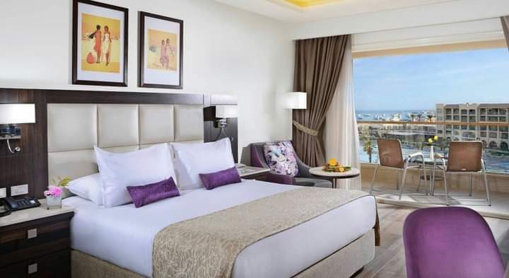 Resort bedroom with white bedding, purple accents, and balcony overlooking coastal town and waterfront buildings.