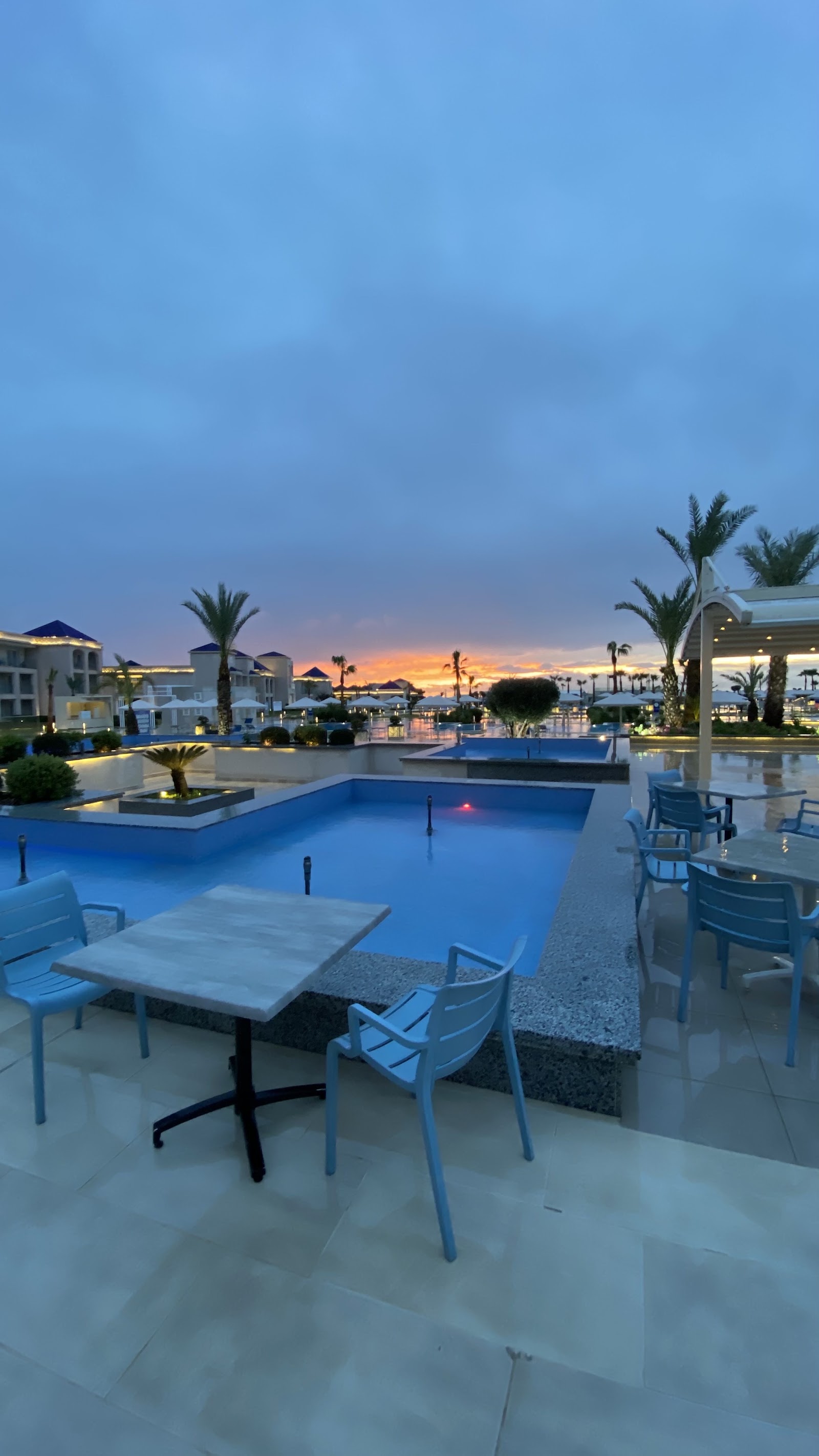 Resort pool and dining terrace at dusk with loungers, palm trees, and orange sunset sky reflected in still water.