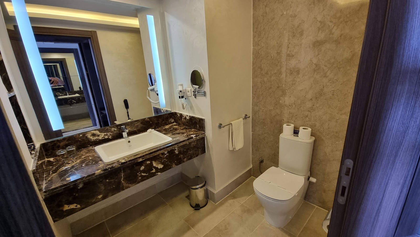Modern bathroom with dark marble vanity, illuminated mirror, white toilet, and tan tile walls at Pickalbatros White Beach Taghazout.