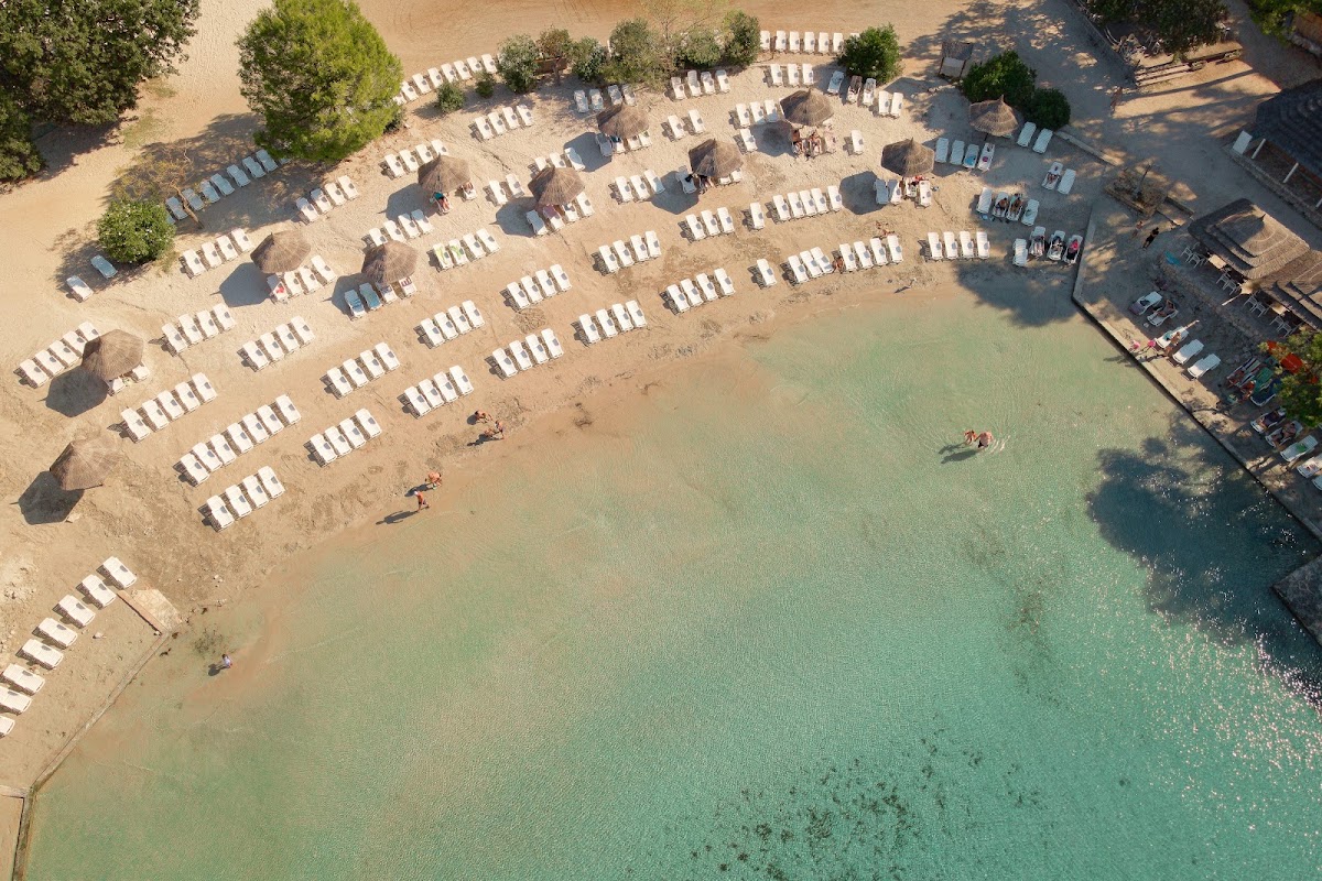 Aerial view of sandy beach with rows of white lounge chairs and thatched umbrellas along the turquoise water at Pine Beach Pakoštane.