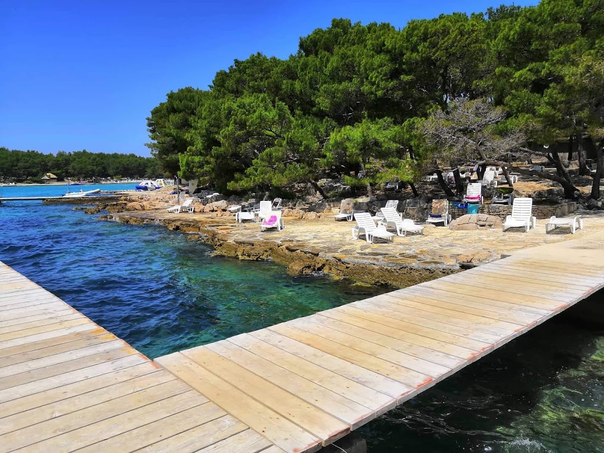 Wooden pier extending into clear turquoise water with white loungers on sandy beach backed by pine trees at Pine Beach Pakoštane.