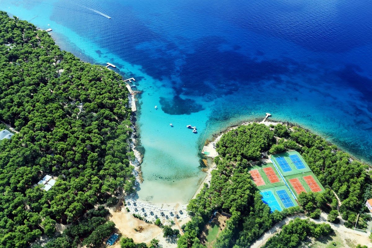 Aerial view of Pine Beach Pakoštane with turquoise waters, sandy beach, tennis courts, and dense green forest surrounding a cove.
