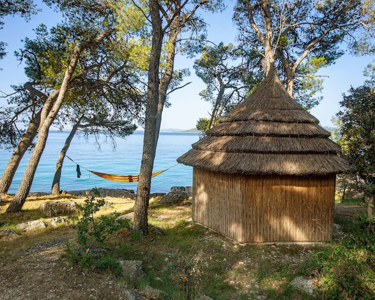 Thatched-roof wooden hut with hammock strung between pine trees overlooking calm blue Adriatic Sea at Pine Beach Pakoštane.