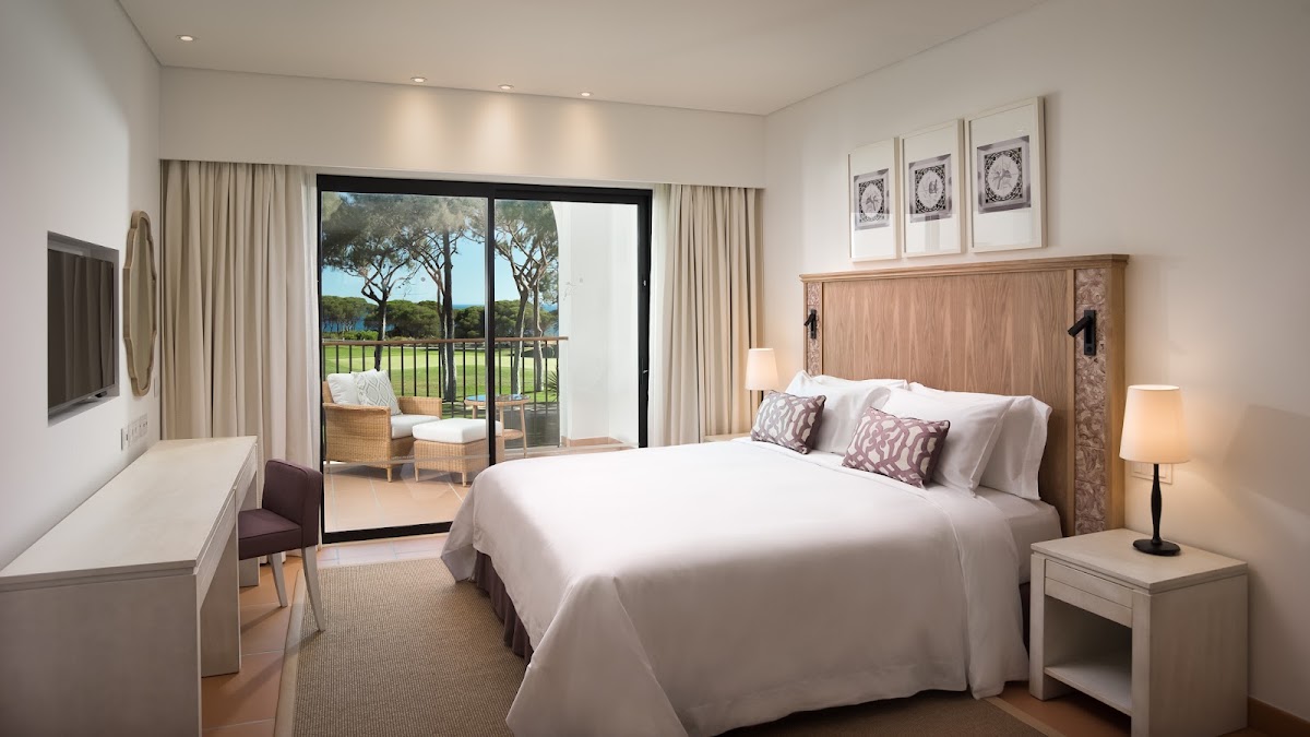 Resort bedroom with king bed, wood headboard, and glass doors opening to a balcony overlooking green grounds and trees.