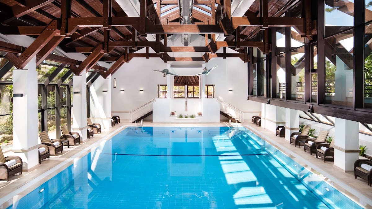 Indoor lap pool at Pine Cliffs Resort with blue water, wooden beam ceiling, lounge chairs on sides, and white walls with large windows.