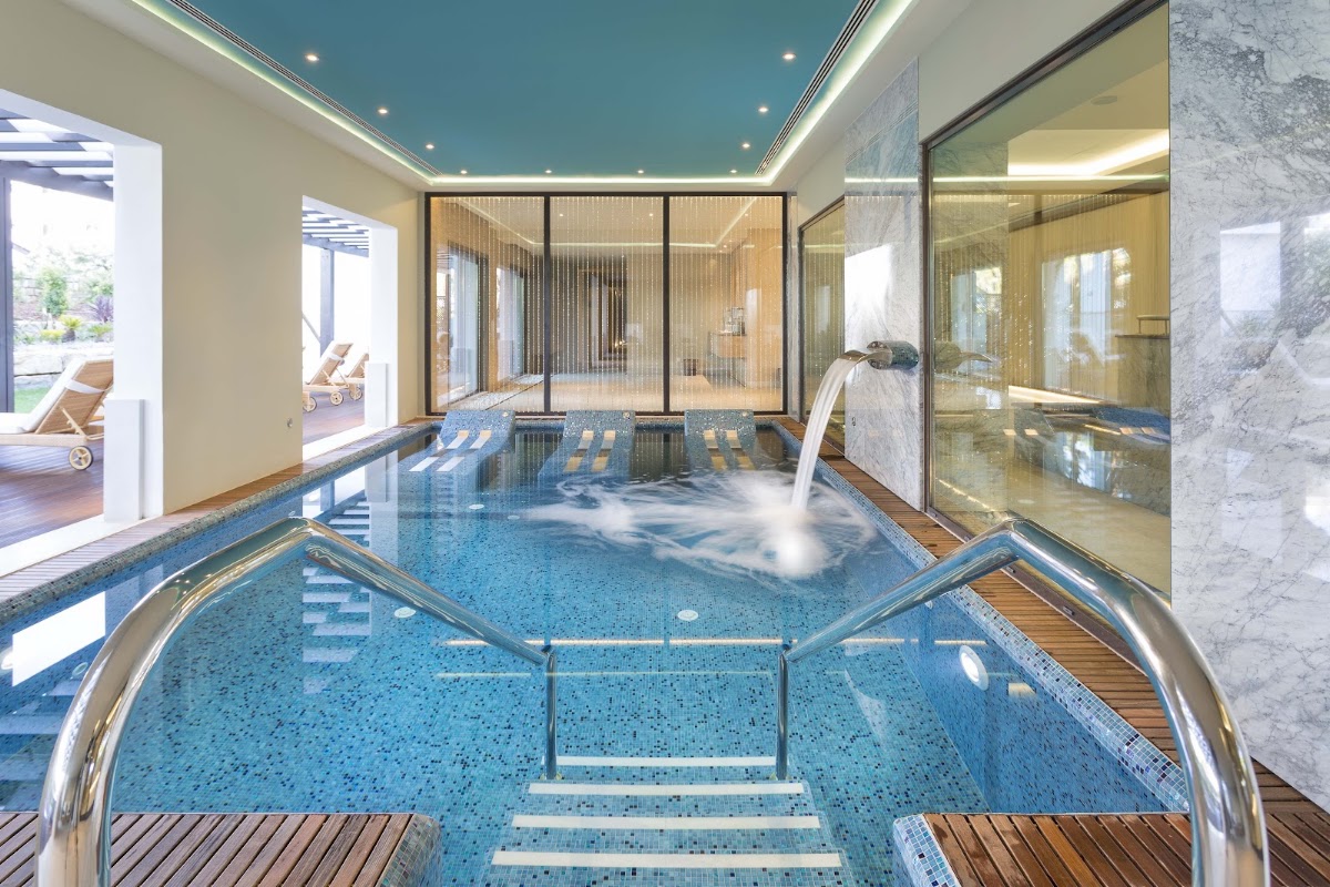 Indoor swimming pool with blue mosaic tiles, metal railings, and a waterfall feature, with glass partitions and access to an outdoor terrace beyond.