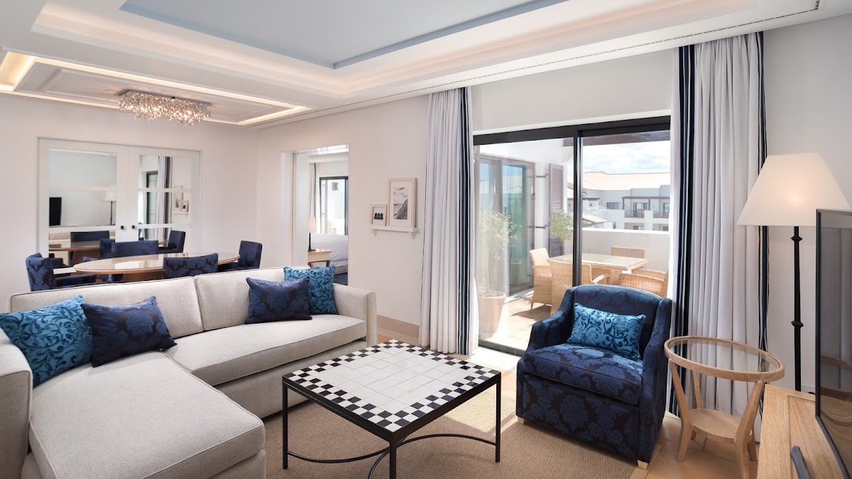 Spacious resort suite with white sectional sofa, blue accent chairs, checkered coffee table, and glass doors opening to a furnished balcony overlooking the grounds.