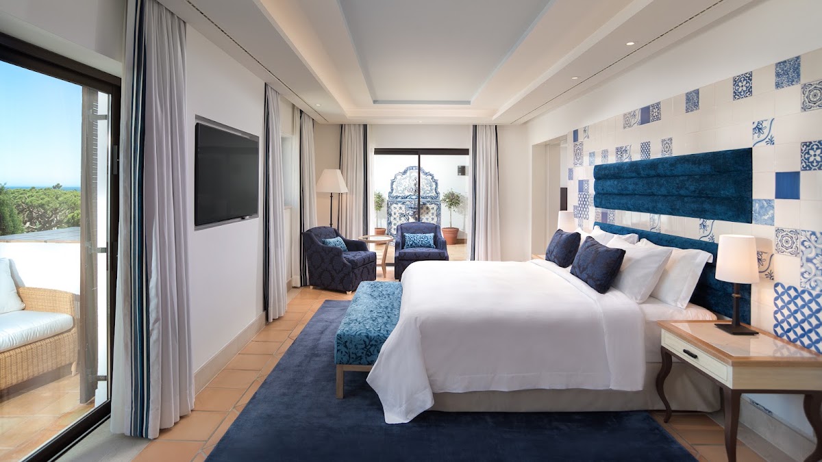 Resort suite bedroom with blue and white tile accent wall, king bed, terracotta flooring, and sea views through glass doors to a terrace.