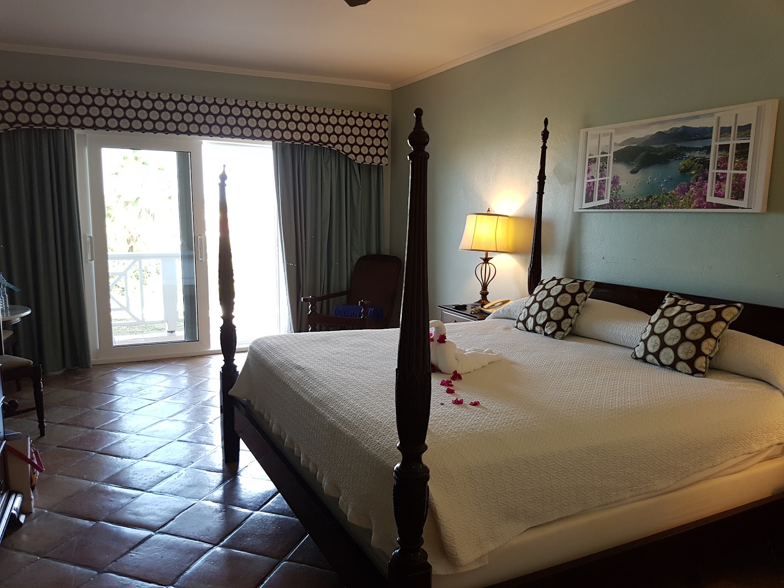 Resort guest room bed decorated with red rose petals and white towel swans, with beach view window art above headboard.