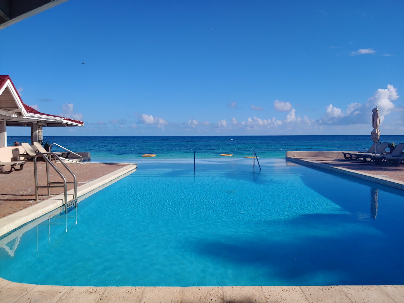 Resort pool with swimmers and loungers overlooking turquoise Caribbean water, palm trees, and beach under blue sky.