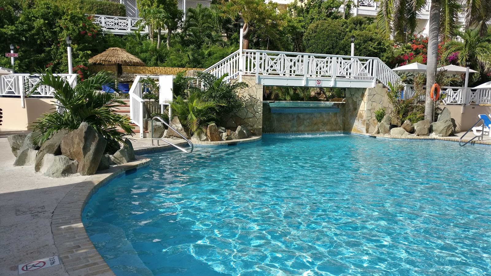 Turquoise pool with rock features and bridge at Pineapple Beach Club Antigua, surrounded by palm trees and white railings.