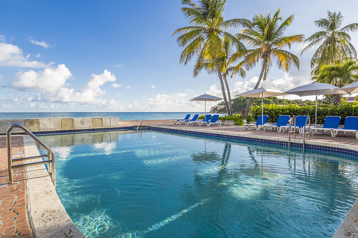 Resort pool overlooking the Caribbean Sea with blue loungers, palm trees, and umbrellas under clear skies.