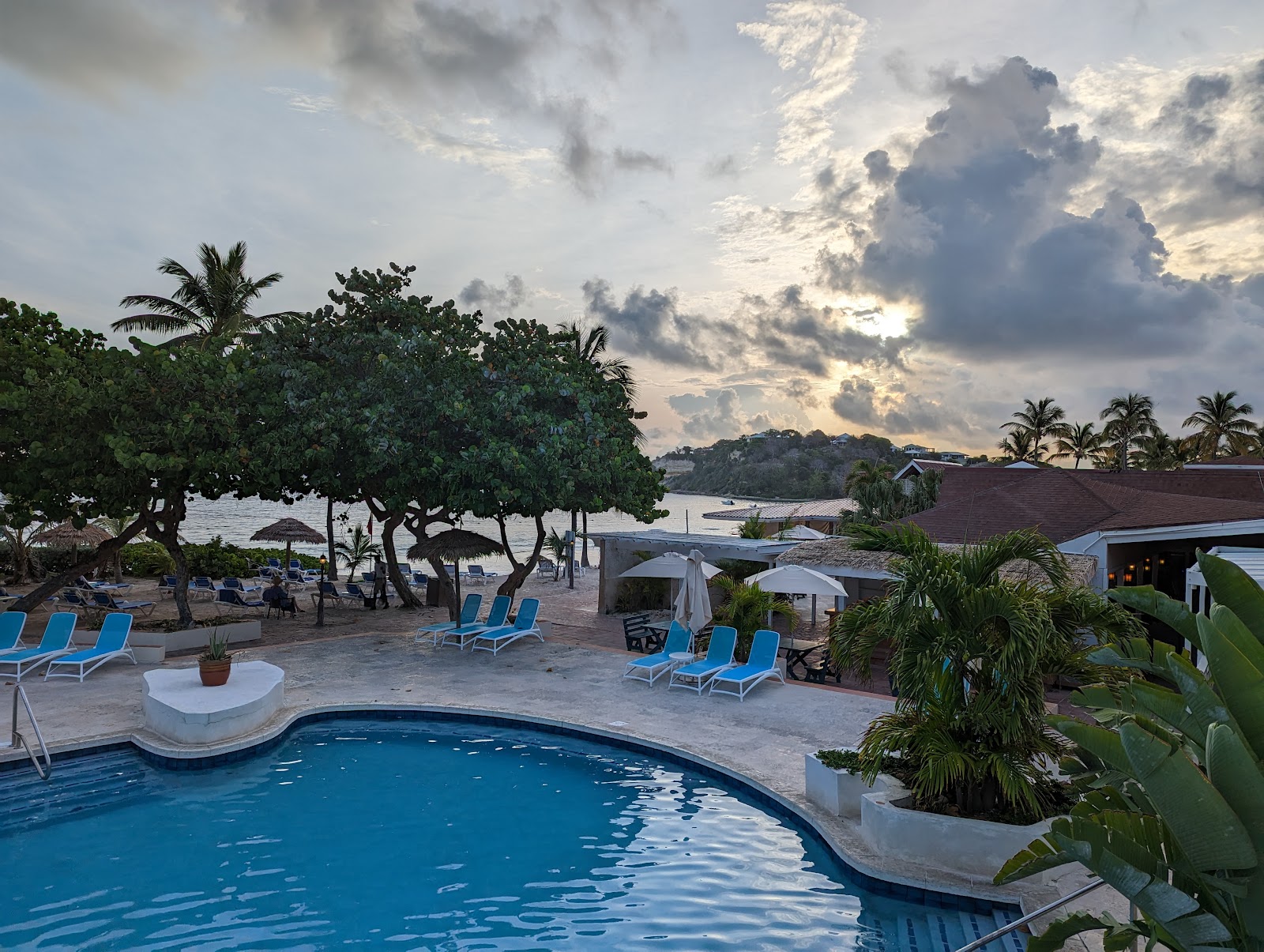 Red-roofed resort buildings overlook a white-sand beach with blue loungers, turquoise waters, and moored boats under a cloudy sky.