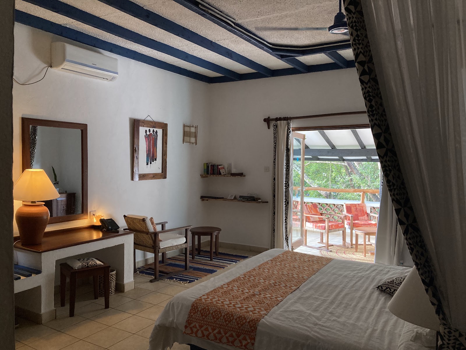 Resort room with striped ceiling, bed with patterned runner, desk with lamp, and glass doors opening to a deck with red chairs and ocean view.
