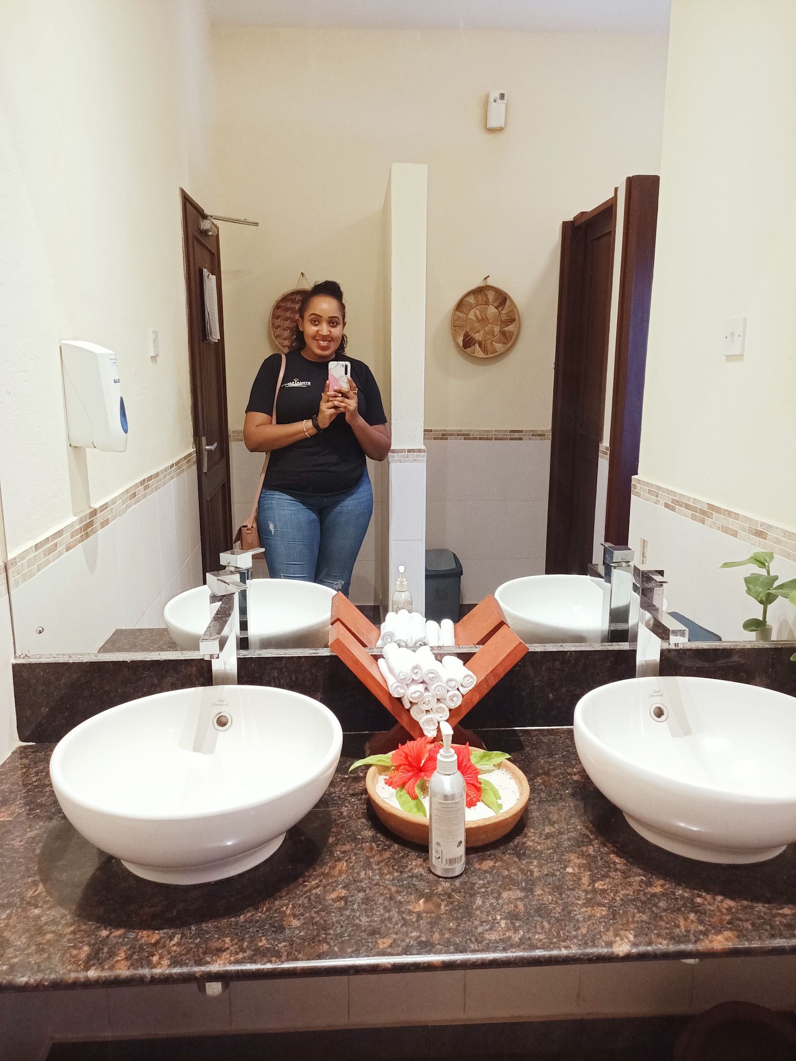 Resort bathroom with dual round white sinks, dark granite countertop, rolled towels, and decorative flowers reflected in large mirror.
