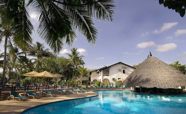 Pinewood Beach Resort's pool terrace with lounge chairs, yellow umbrellas, and thatched-roof structures surrounded by palm trees under blue sky.