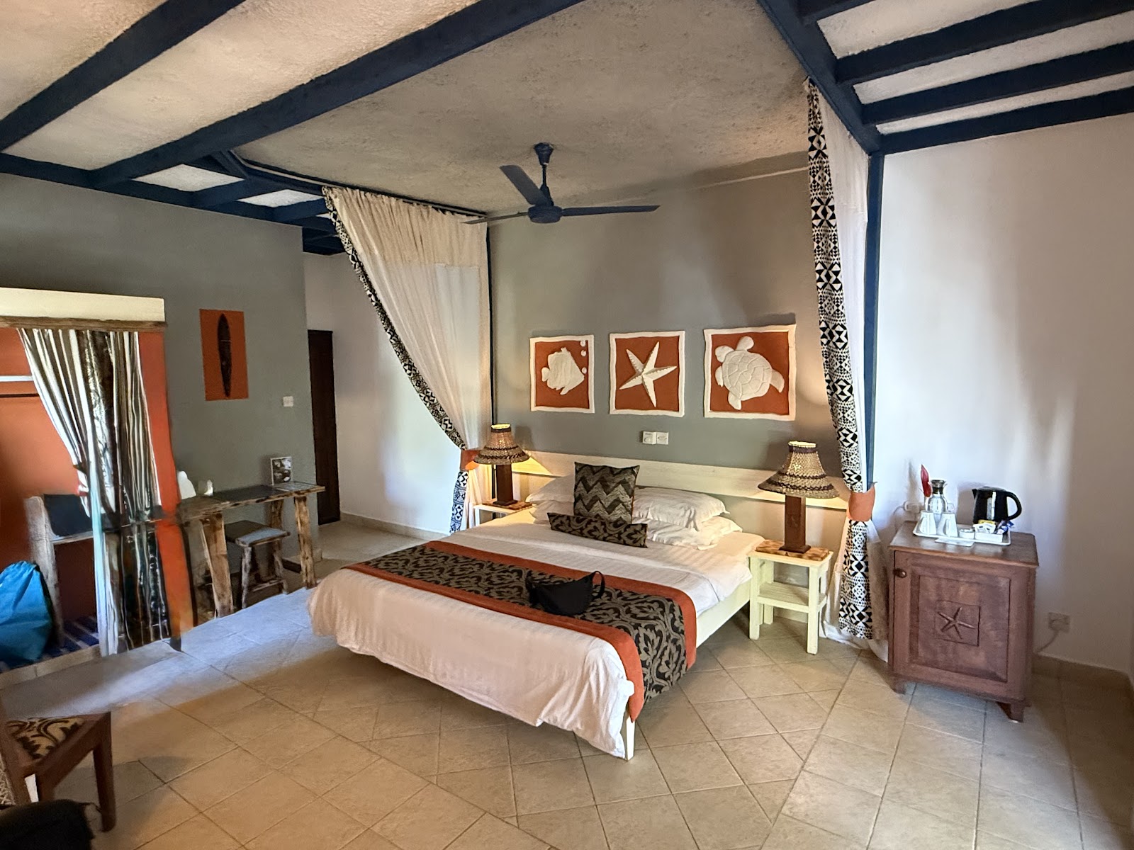 Resort bedroom with four-poster bed, ocean-themed wall art, tropical furnishings, and open-air design with ceiling fan.